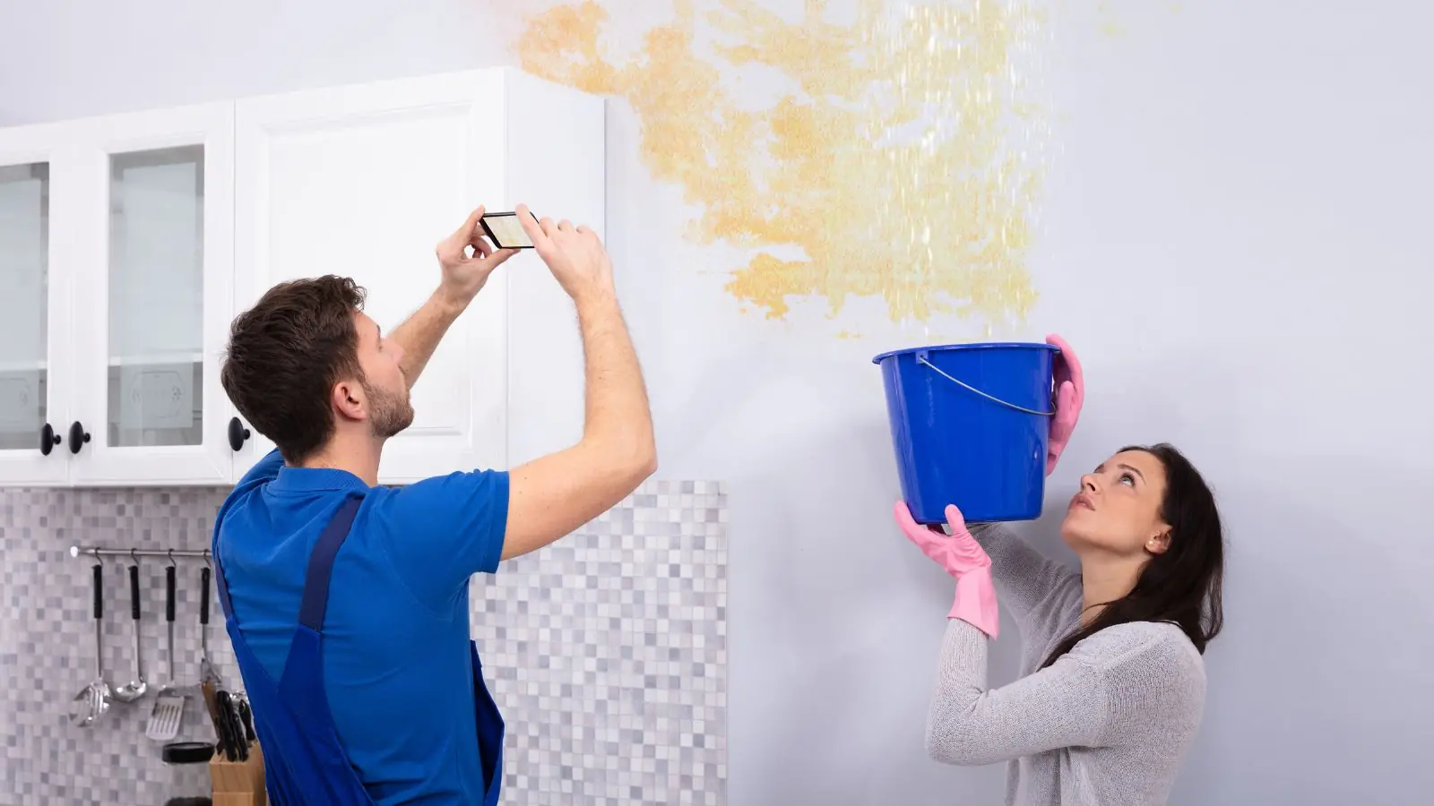 Renter photographing a water stained wall while another holds a bucket under a leak